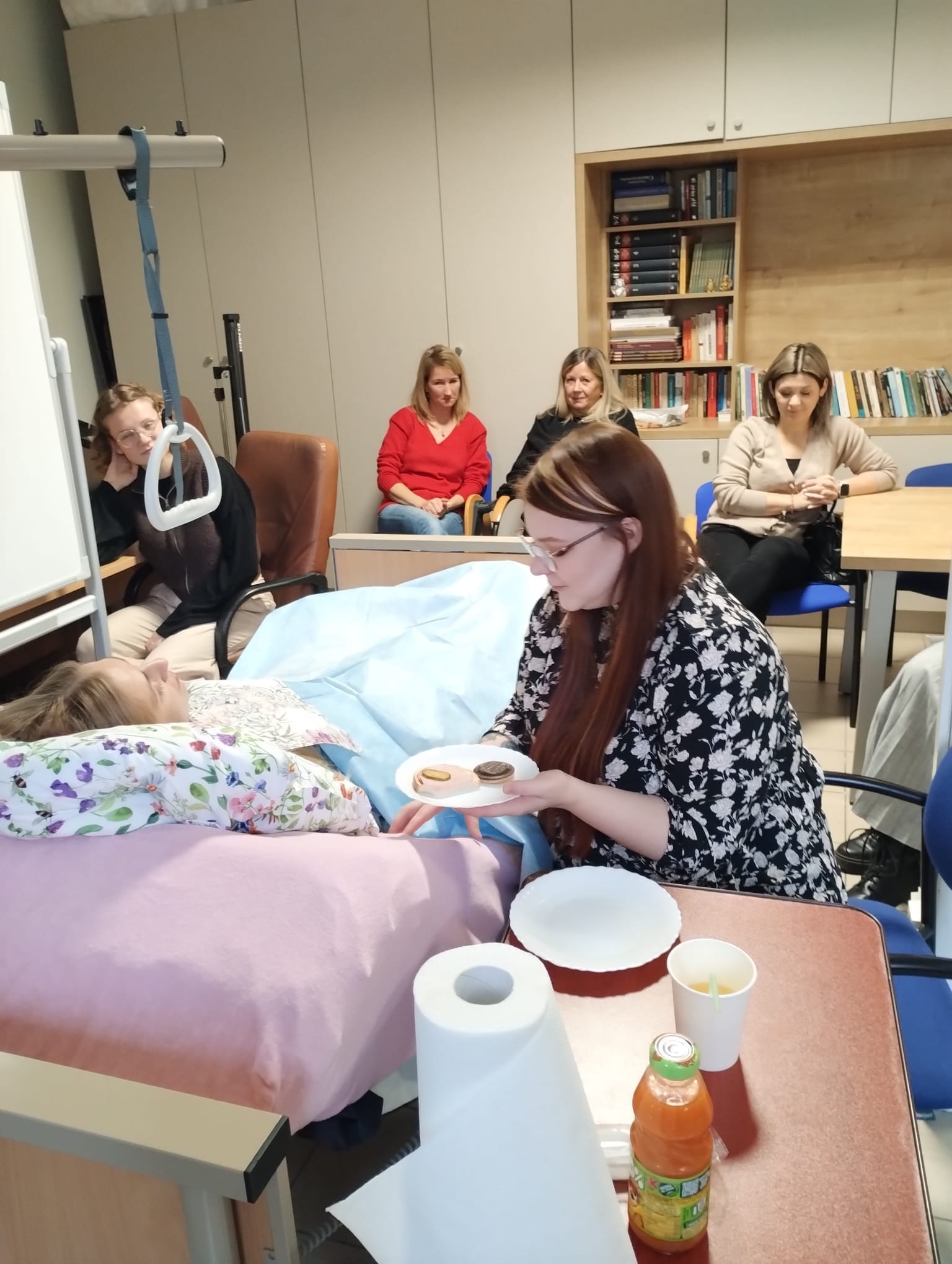 wolontariuszka karmi osobę leżącą na łóżku. Kilka innych osób przygląda się tej czynności.