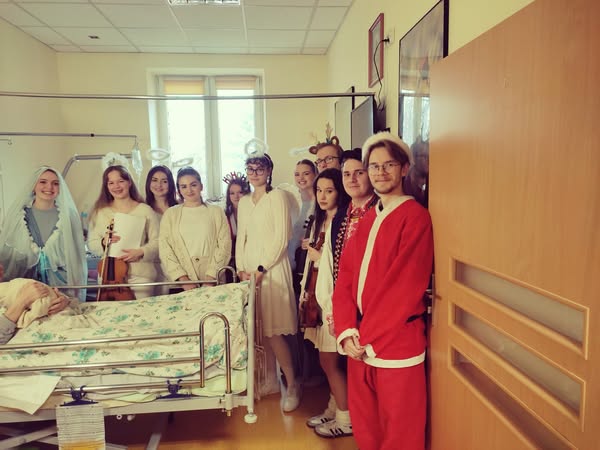 Grupa wolontariuszy w strojach świątecznych stoi w pokoju szpitalnym przy łóżku chorej
