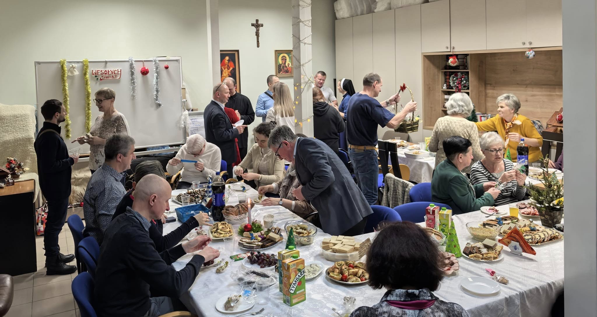 Grupa kilkunastu osób w pomieszczeniu. Część osób stoi, inne siedzą przy świątecznie zastawionych stołach