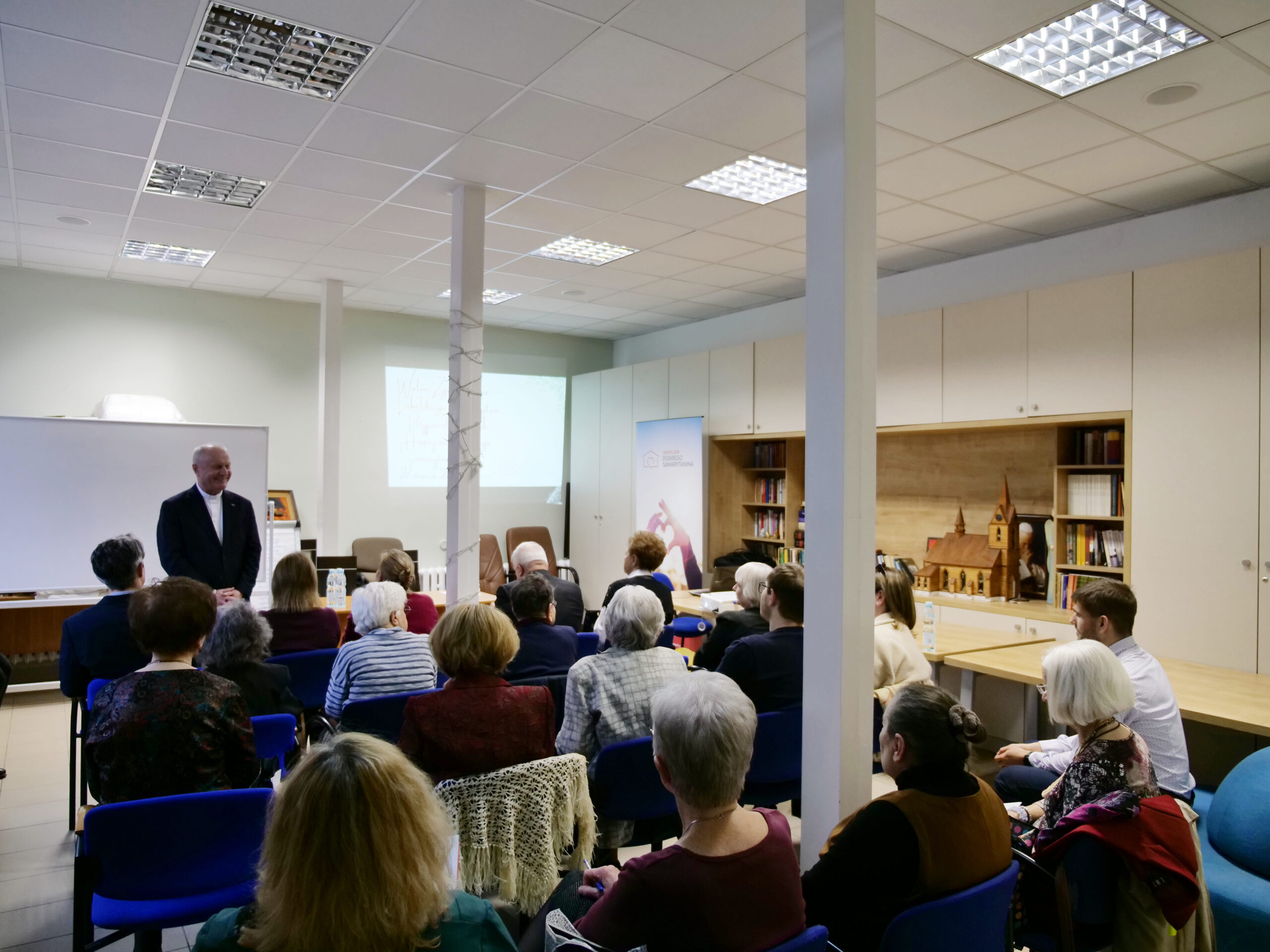 Kilkanaście siedzących osób spogląda na stojącego przed nimi mężczyznę